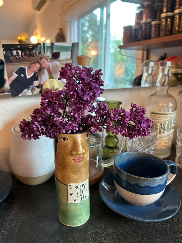 Photo of a green vase with a brown woman's face on it that says "Love will win" holding lilacs, a blue mug, a white vase, some clear glass cups and candlestick, and a photo of me and my mom