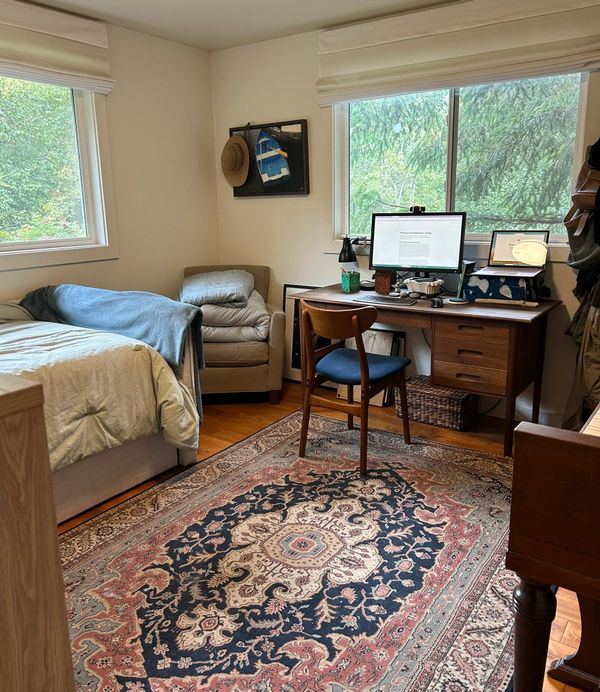 A photo of a home office with a view of Douglas fir trees, a wood desk, an old dark blue and rose rug, the corner of a piano, a gray comfortable chair, and a single bed for napping