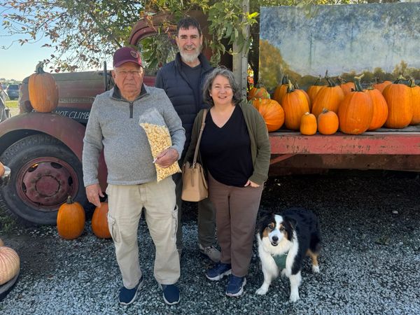 A photo of our family at a pumpkin patch, surrounded by bright orange pumpkins, a rusty old truck, blue sky, a tree turning red, and our a smiling fluffy Australian Shepherd dog.