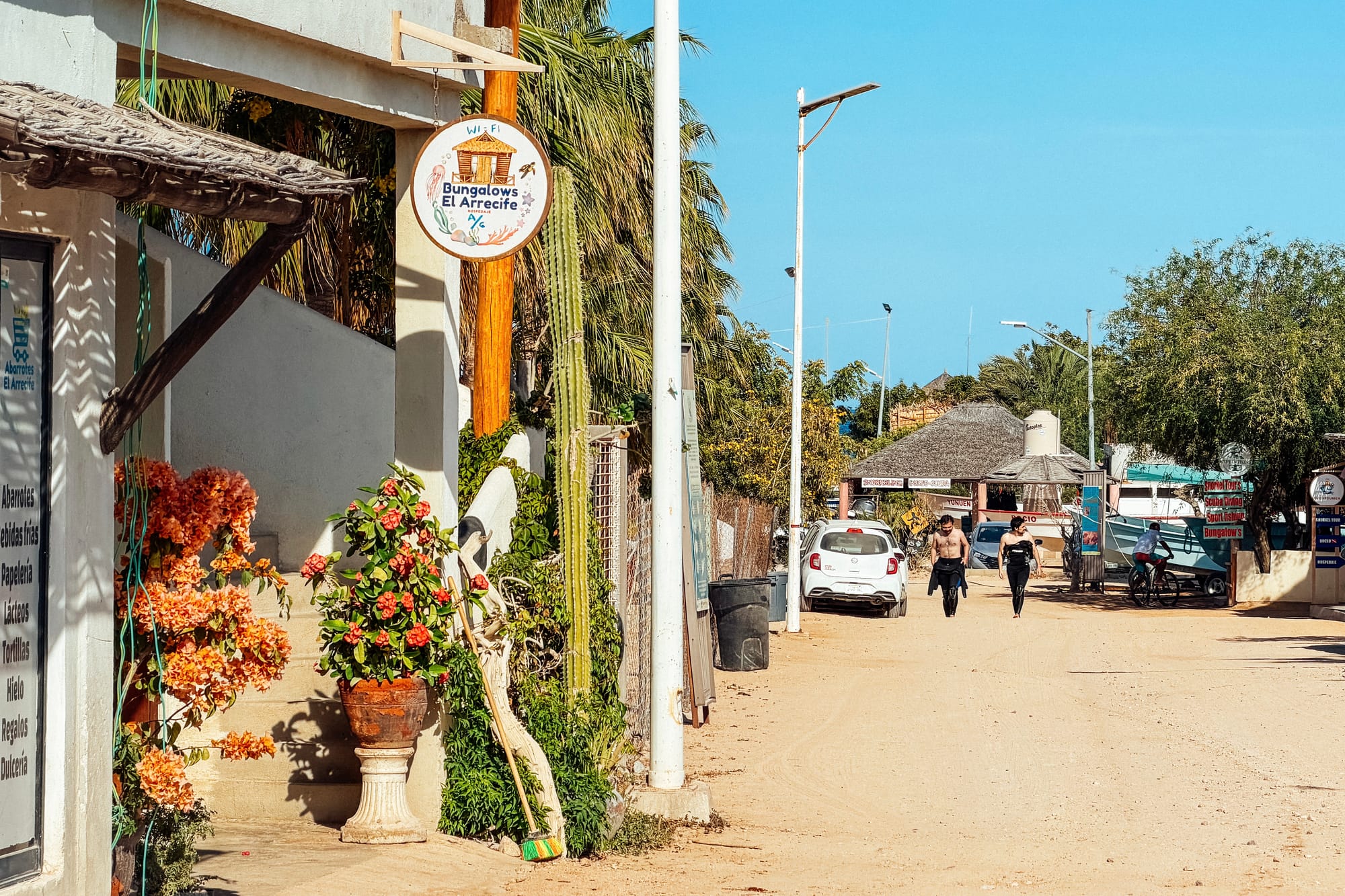 Strada polverosa nel centro di Cabo Pulmo con centri immersioni e due subacquei in muta che camminano verso il villaggio.