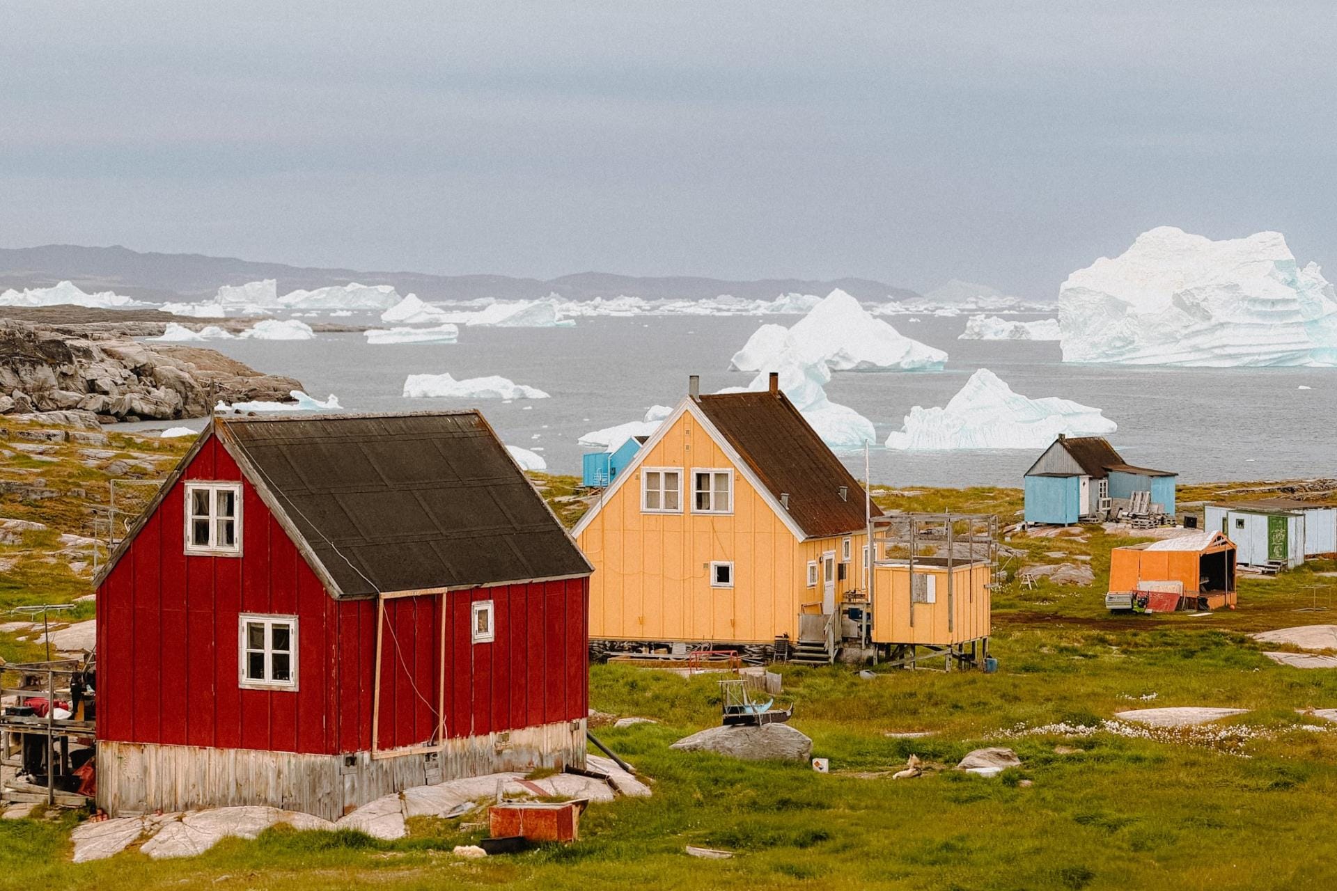 Case di legno rosse, gialle e azzurre della comunità di Oqaatsut sulla costa della Groenlandia occidentale, con iceberg nella baia sullo sfondo.