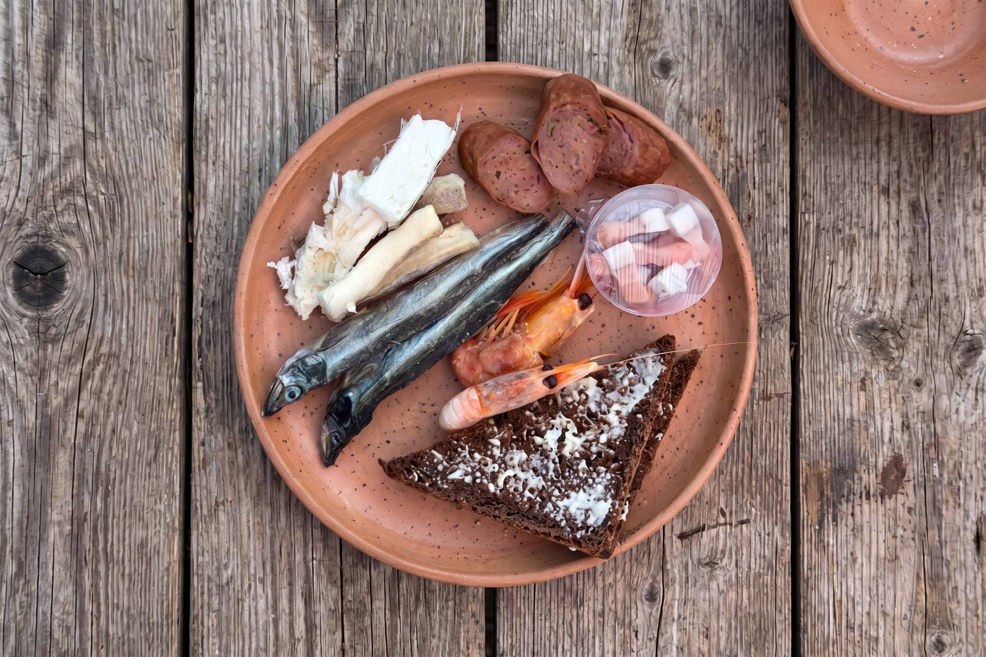 Piatto di terracotta visto dall’alto su assi grigie, con kaffemik groenlandese: pesce secco, salsiccia, gamberi, pane di segale e una tazzina.
