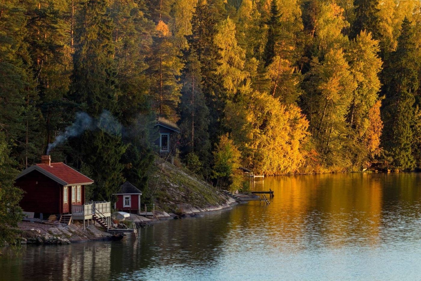 Sauna di legno rossa sulla riva di un lago tra foreste dai colori autunnali in Finlandia, con fumo dal camino