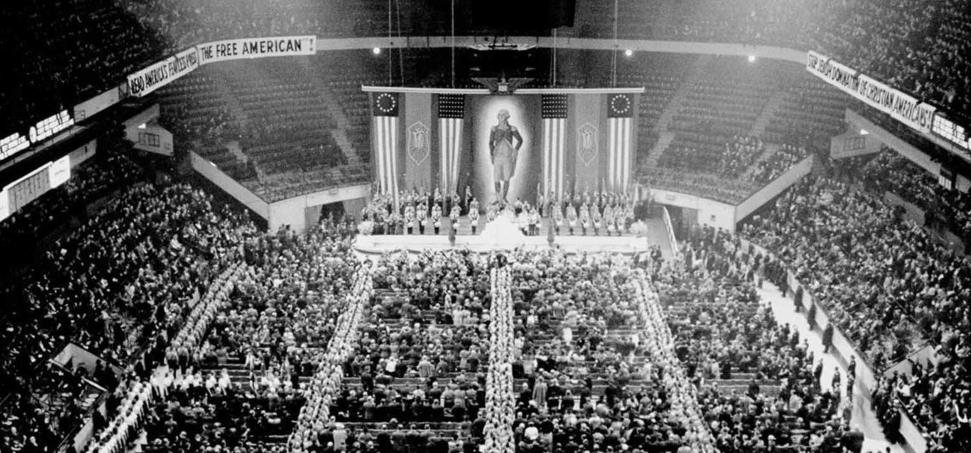 A crowded auditorium hosts a large gathering with a central stage flanked by American flags. Banners read, “Save America” and “The Free American.” It is a pro-Nazi party rally. The mood is solemn and formal.