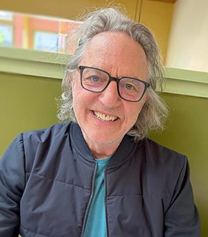 Smiling man with glasses and gray hair, in a casual blue shirt and dark jacket, sitting indoors against a green background. Warm, cheerful atmosphere.