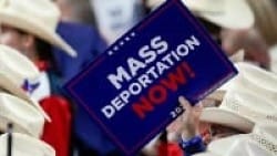 A person in a crowd holds a sign that reads "Mass Deportation Now!" in bold, white and red letters against a blue background. Many wear white cowboy hats.