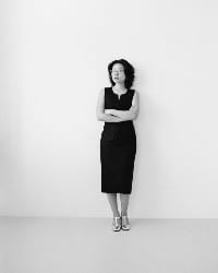 A woman stands against a white wall, in a knee-length black dress and silver shoes. She crosses her arms and gazes thoughtfully into the distance.