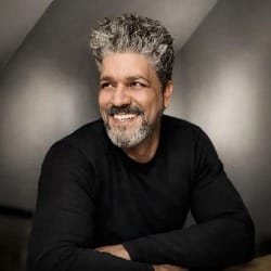 A smiling man with curly gray hair and a beard is sitting with folded arms, wearing a black sweater. The background is softly lit, creating a warm, relaxed atmosphere.