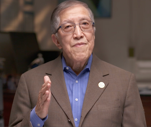 Photo of Dr. Derald Wing Sue, an elderly Asian man wearing glasses, a brown blazer, blue shirt, and lapel pin, gesturing with a raised hand and warm expression.