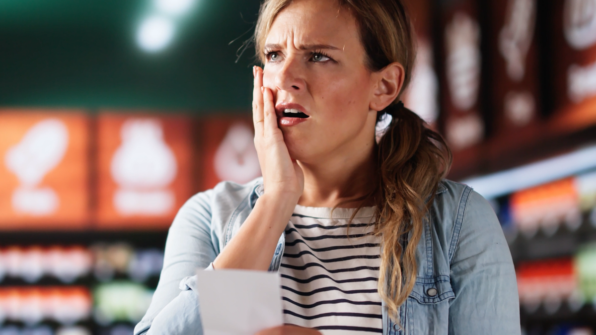 A worried woman in a grocery store presses one hand to her cheek while holding a receipt, her furrowed brow and open mouth showing alarm.