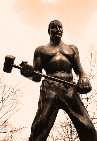 Bronze statue of a muscular man - John Henry - holding a sledgehammer, facing forward. Leafless trees and a cloudy sky form the background, creating a solemn tone.