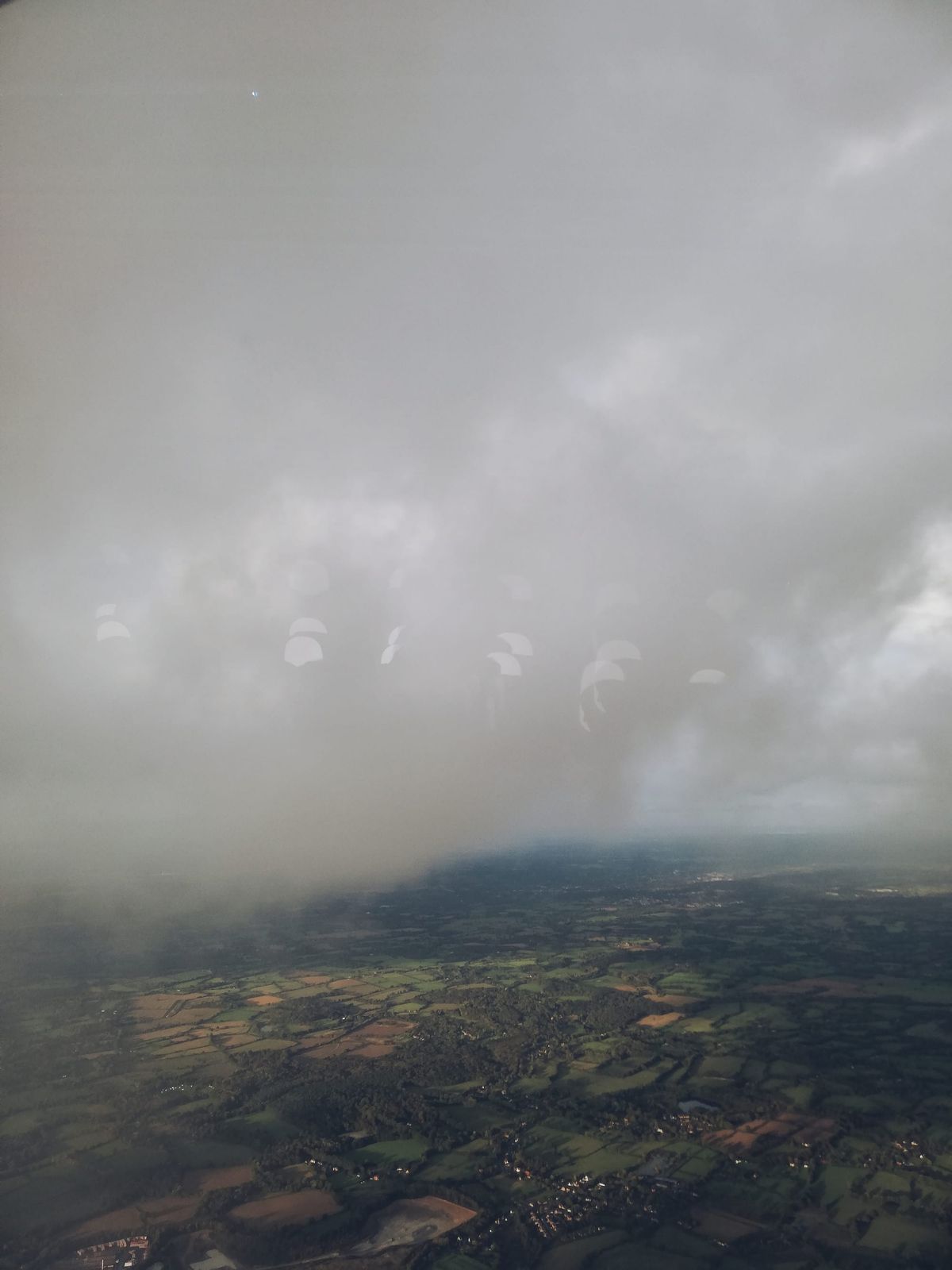 foto tomada desde la ventana de un avión donde se ven prácticamente solo nubes espesas y grises y un terreno abajo mayoritariamente de un verde bosque
