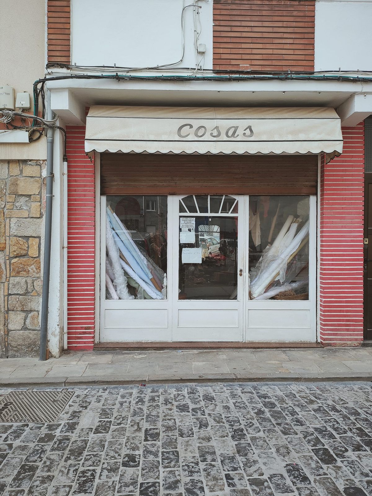 una tienda aparentemente cerrada hace mucho tiempo con rollos de tela dentro y un toldo exterior que dice "Cosas"