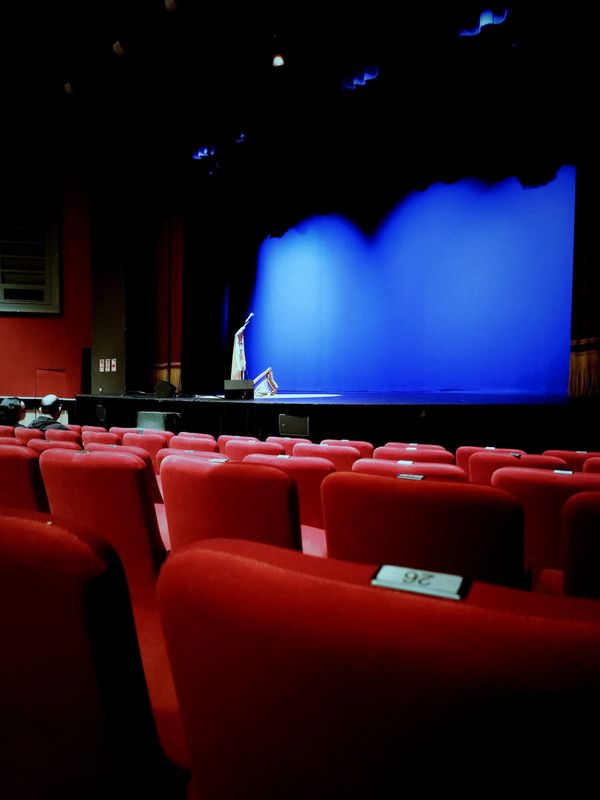 unas butacas vacías de teatro, color rojo y un escenario con pantalla azul