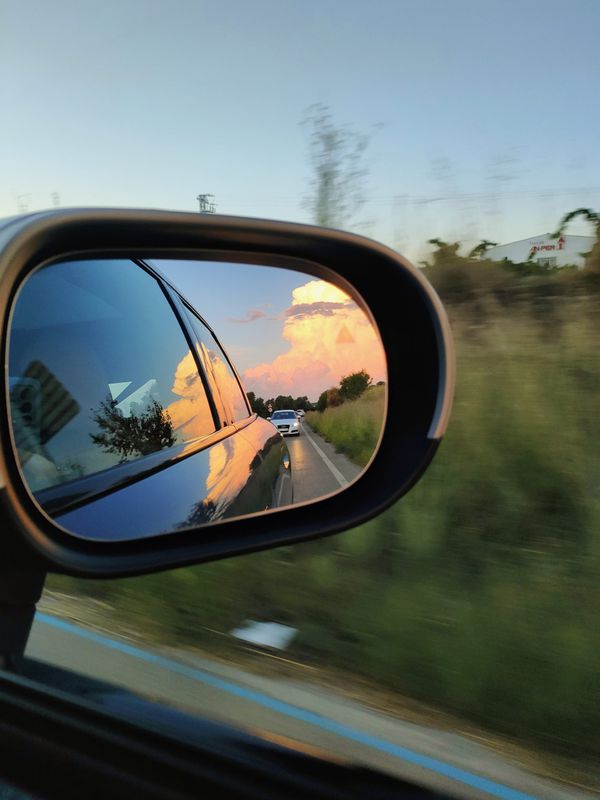 una foto tomada desde dentro de la ventanilla cerrada de un coche que enfoca una nube muy grande en el atardecer, reflejada en el retrovisor del coche