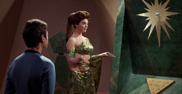 McCoy kneels before an altar with a sun symbol. Between them, a priestess holds a small electronic device.