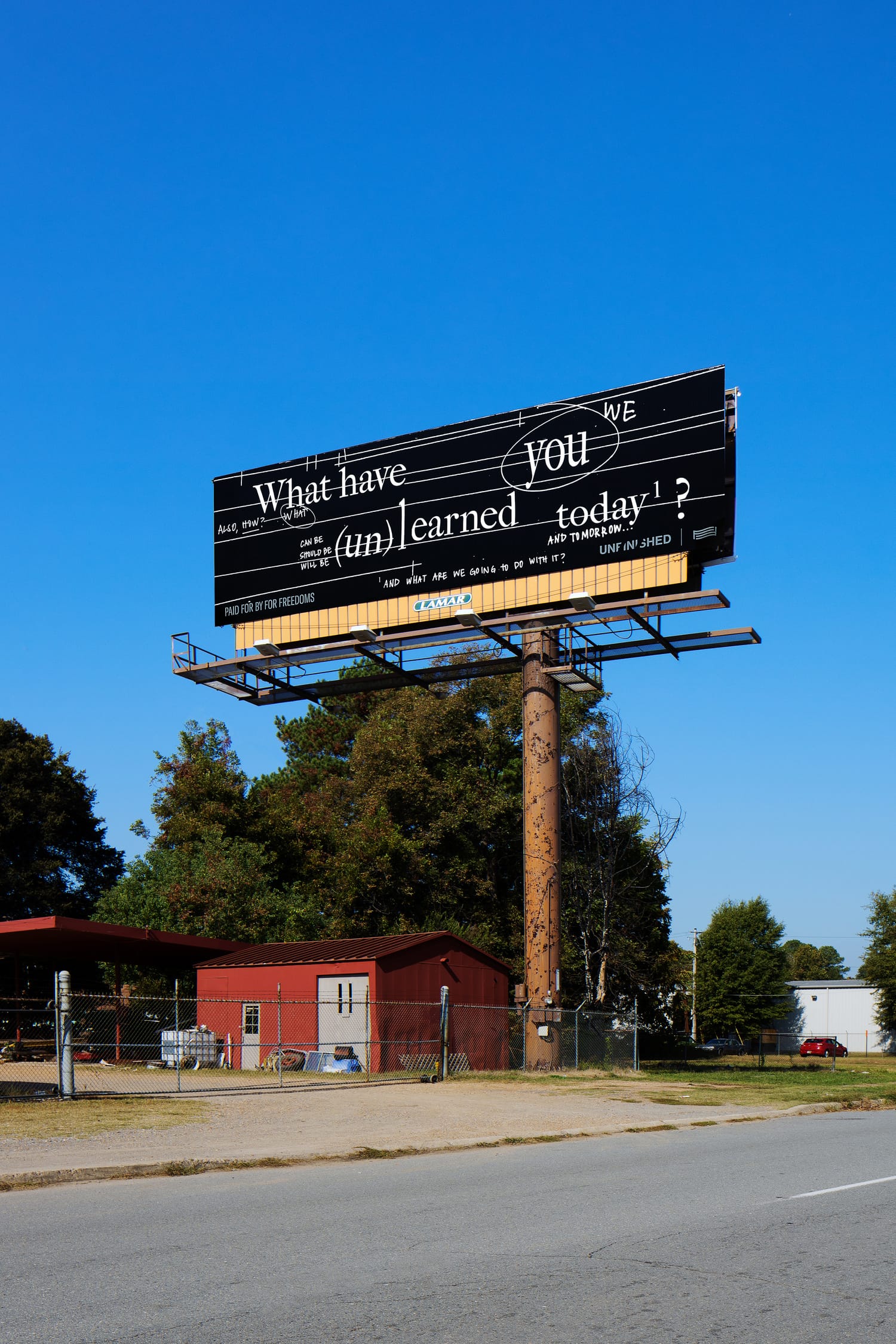 Photo of an elevated black billboard by an American suburban road with a white stylised slogan “What have you unlearned today?”