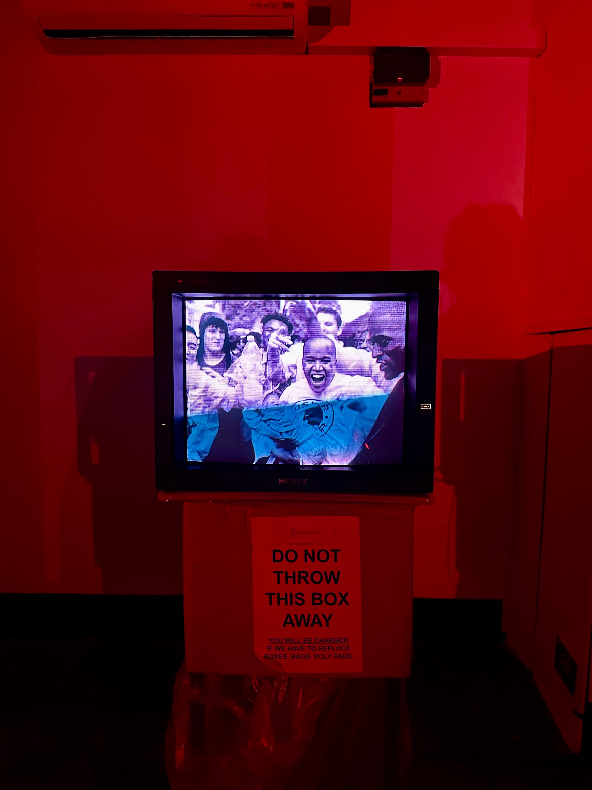 A CRT TV showing black people at carnival in a dimly lit gallery bathed in red light.