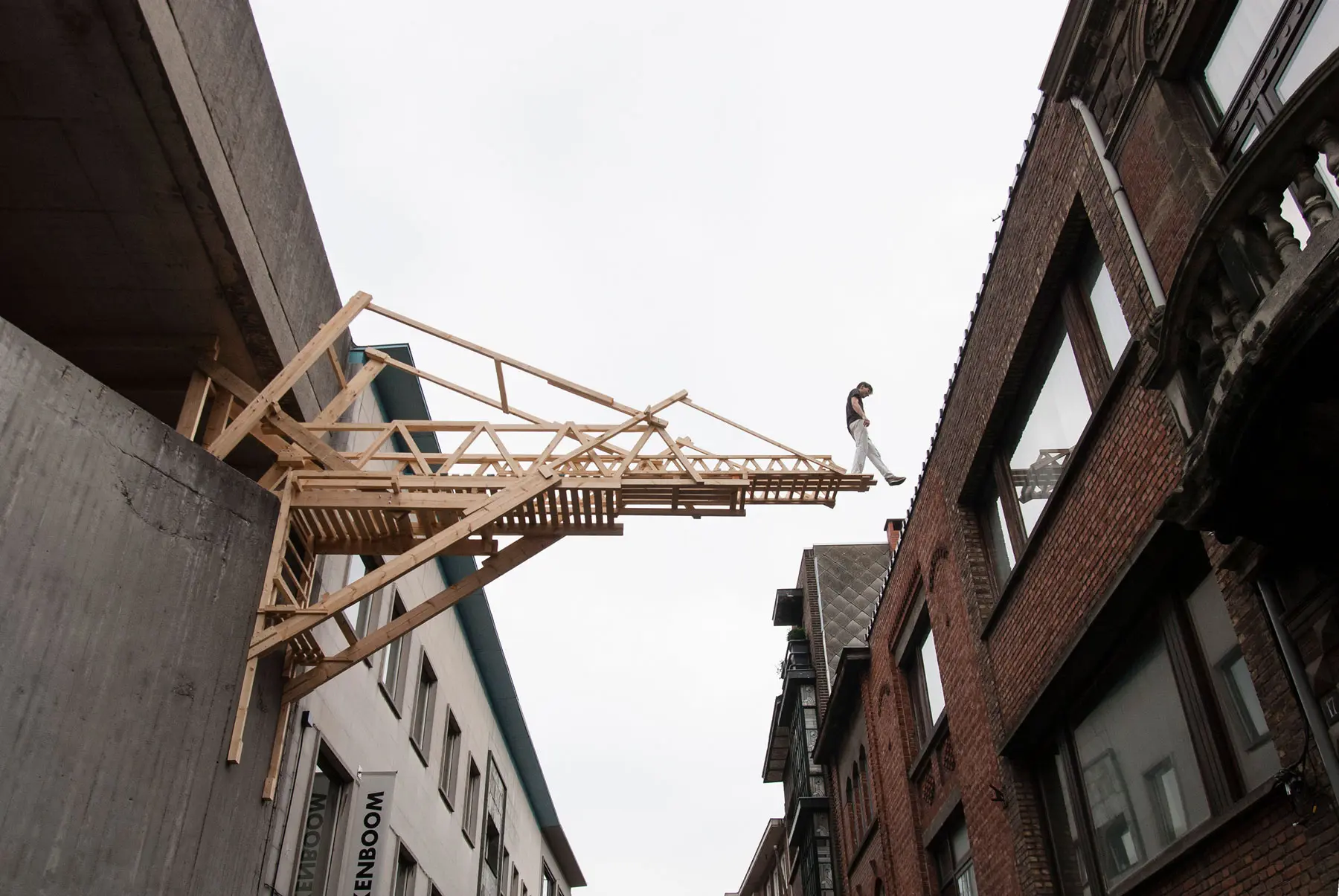 Photo taken from street level of a wooden bridge structure between two buildings, with a person walking across appearing to step over its edge.