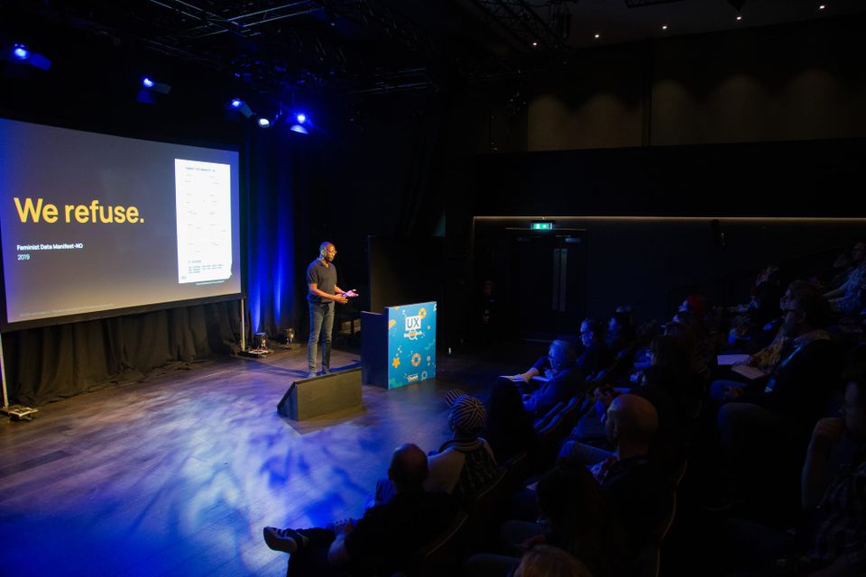 A tall Black man in a navy short sleeved polo shirt and dark jeans speaking to an audience illuminated by blue light.