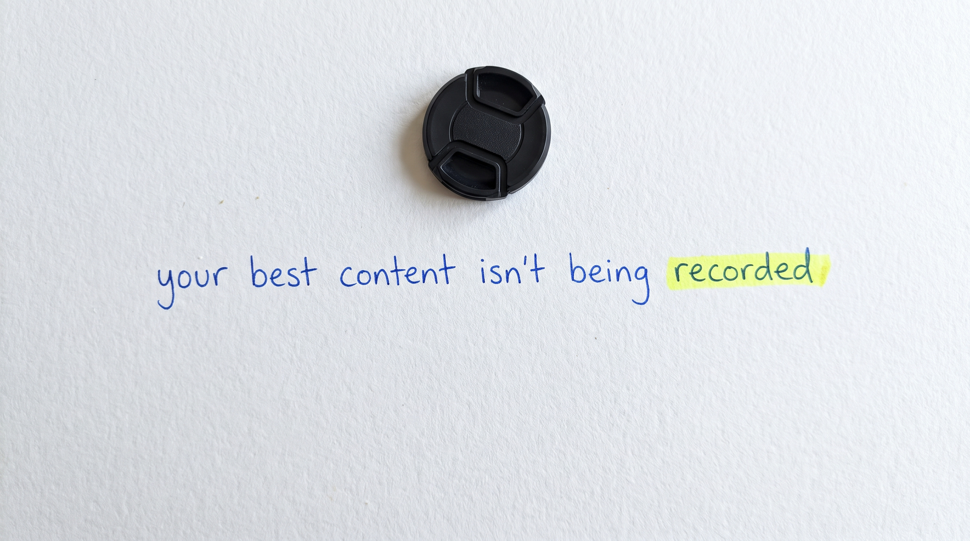 Handwritten blue pen on white paper reading "your best content isn't being recorded", with a camera lens cap lying flat above it