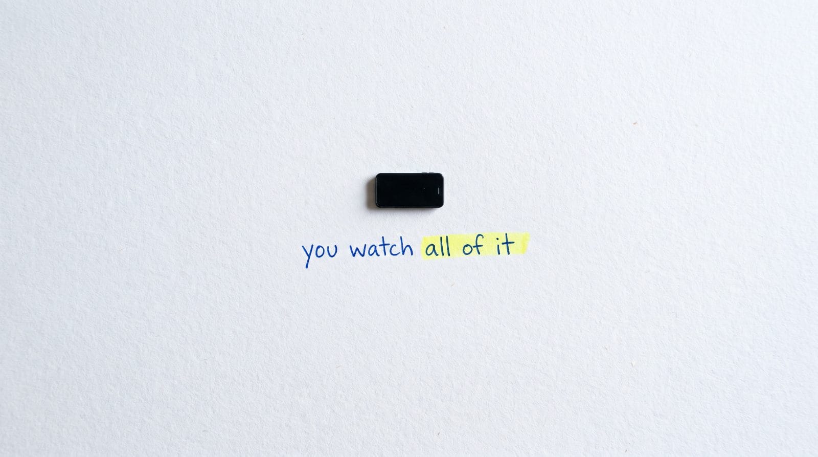 A miniature phone on white paper with the handwritten words: you watch all of it. The word all is highlighted.