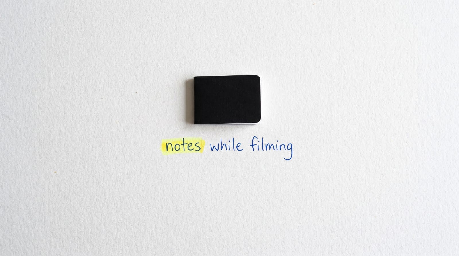 A closed black pocket notebook on white paper with the handwritten words: notes while filming. The word notes is highlighted.