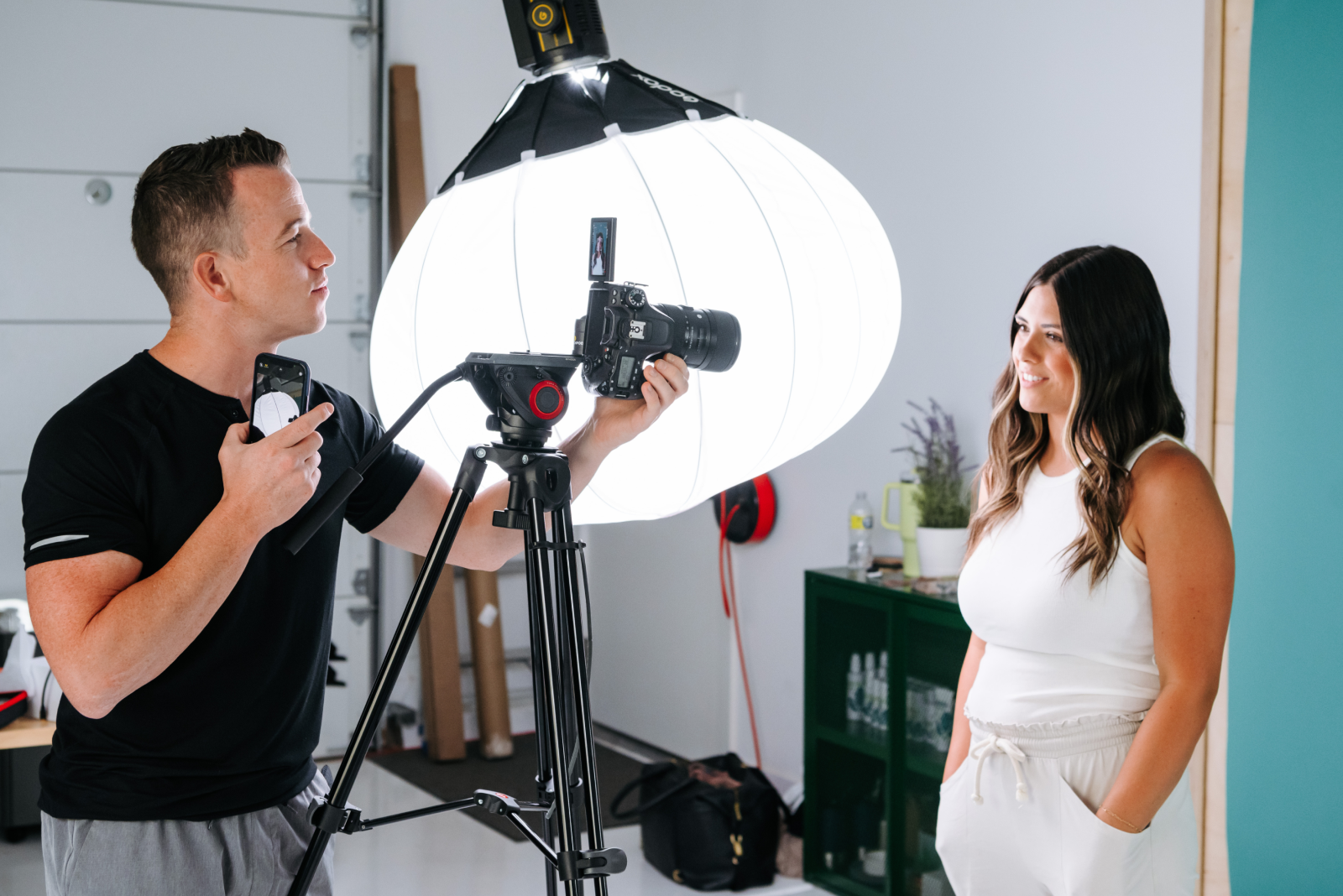 A full studio setup with a large softbox, camera on tripod, boom mic and branded backdrop, one person seated at a table