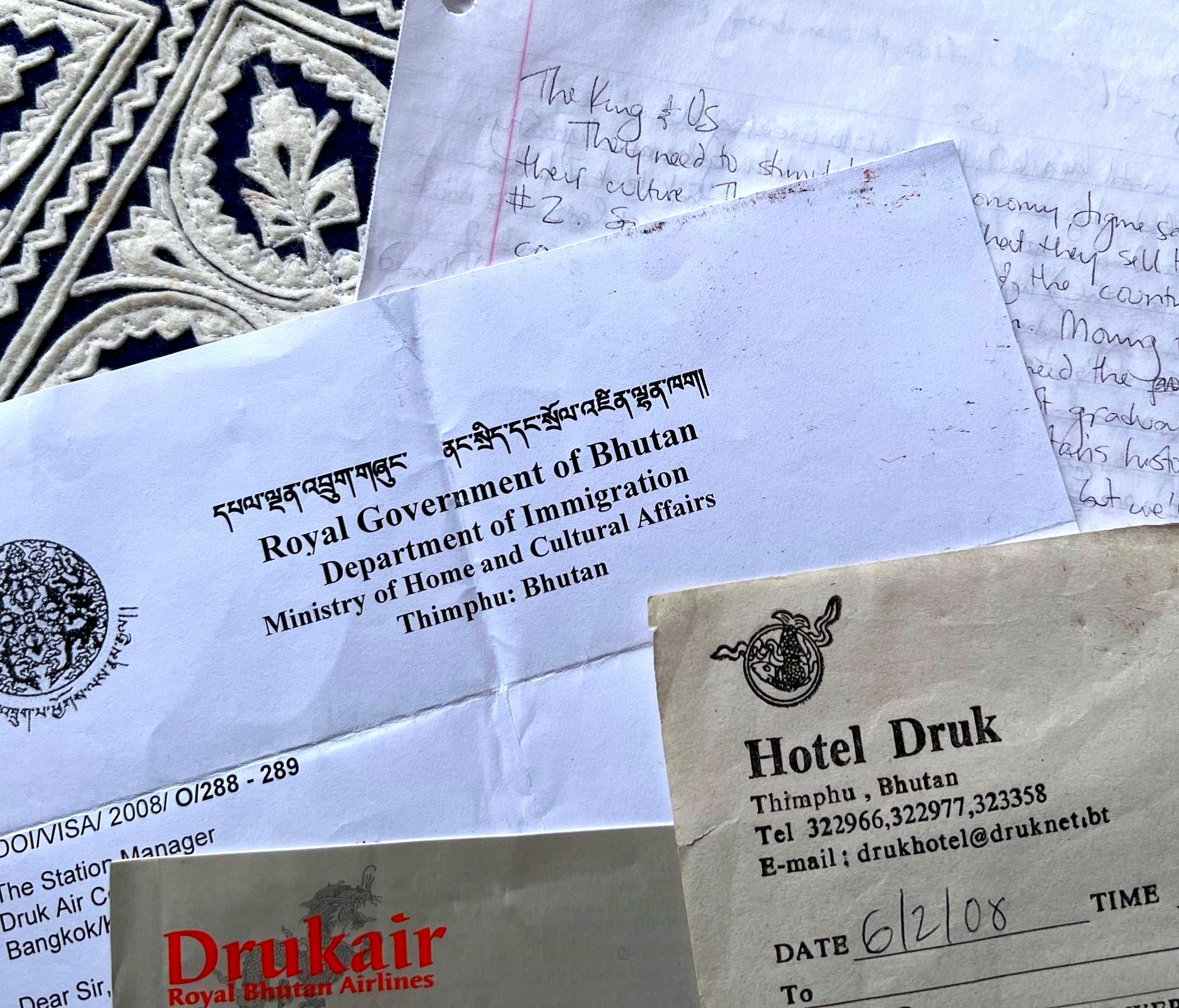 Four documents arrayed against a background of royal-blue-and-white fabric in the lefthand corner. The top right document looks like handwriting on notebook paper, with the heading “The King and Us”; below and to the left of that is a printed document that headed with “Royal Government in Bhutan Department of Immigration” in both Bhutanese and English and appears to be a visa; to the bottom and right of that, a paper headed with the words “Hotel Drunk” and contact information in Thimpu, Bhutan, and the handwritten date 6/02/08; at the bottom of the photo is a darker brown piece of paper that says “Drukair, Royal Bhutan Airlines” in orange text.