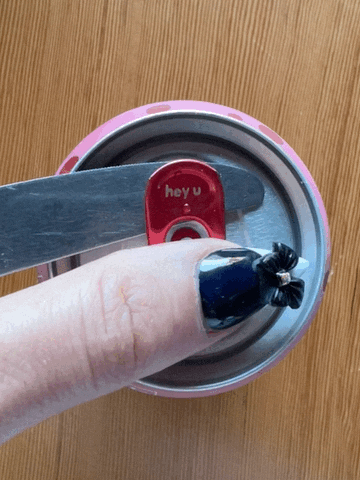 A tab on an aluminum can opened between a butter knife and a long black stiletto thumbnail with white tip.