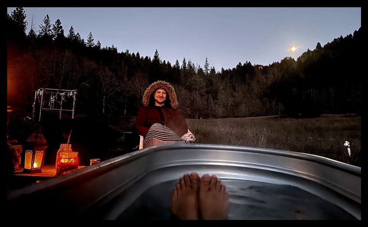 Moon hovering above a pine-covered hill behind the author. Foreground is a pair of feet poking out of a h