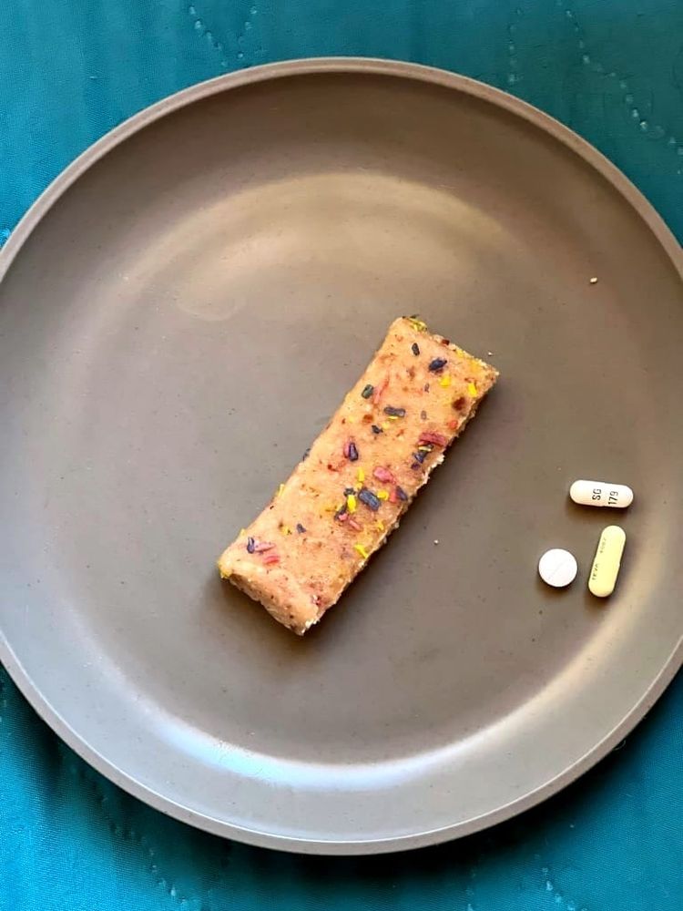 A small rectangle of cashew-colored confection with sprinkles on a grey plate with 3 pills.