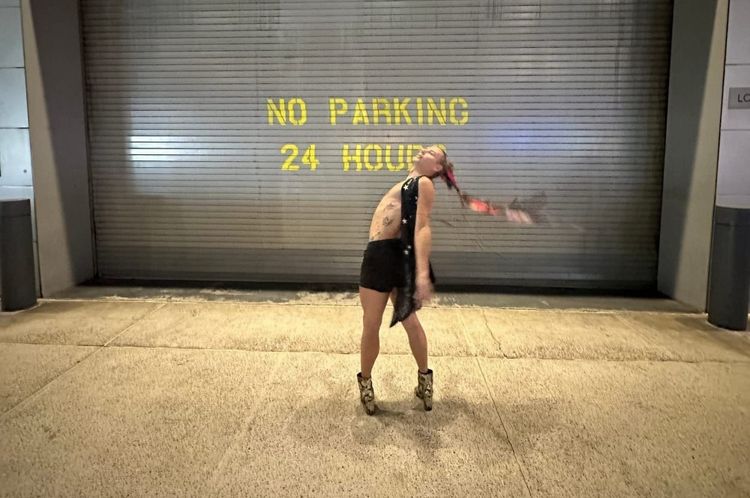 A person in high-heeled alligator boots, shorts, and a sequin vest stands in front of a garage painted NO PARKING.