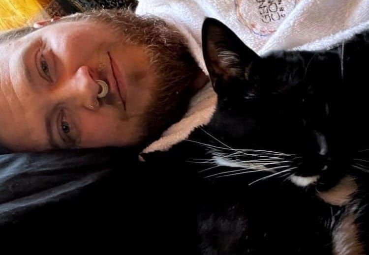 A man with a beard lying next to a sleeping black cat with white whiskers.