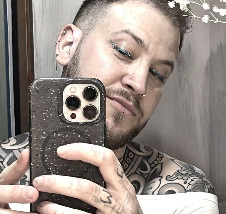 A man with a beard and blue-glitter cat-eye eyeliner takes a selfie in a mirror. He has black, flowery tattoos covering his bare shoulders.