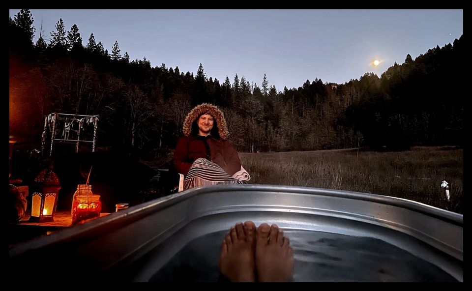 Moon hovering above a pine-covered hill behind the author. Foreground is a pair of feet poking out of a h