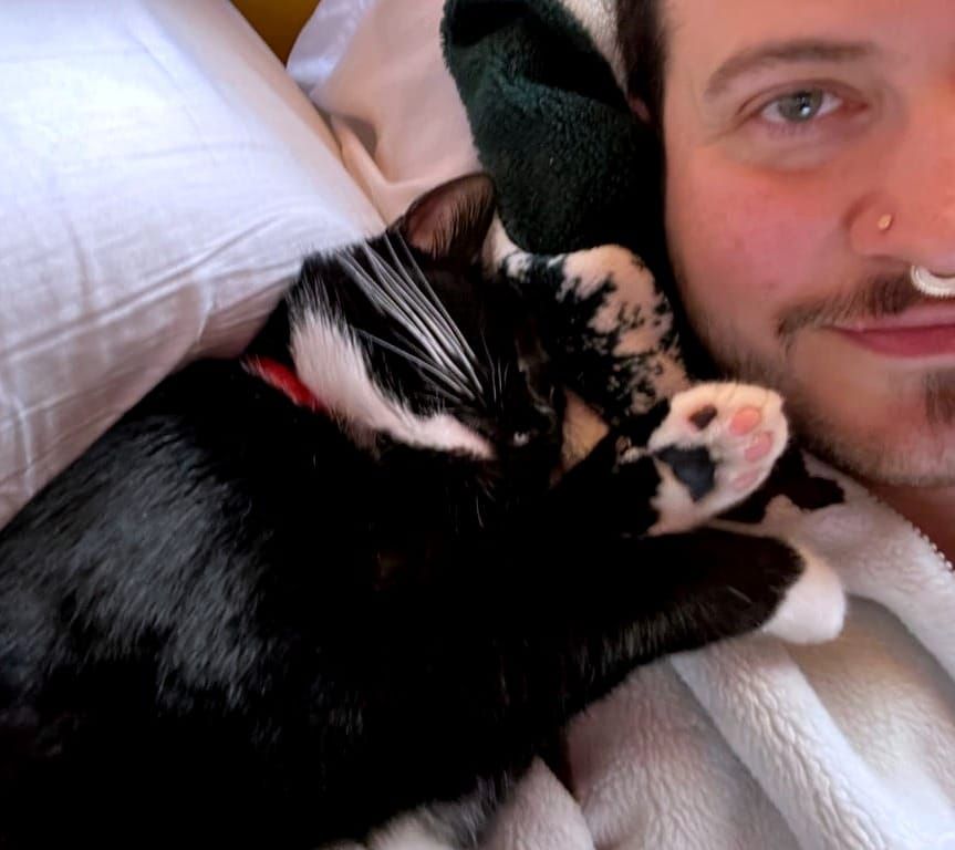 A man lying in bed with a cat asleep on his shoulder. The cat is black with white paws and whiskers; the man is white and wearing a white fleece.