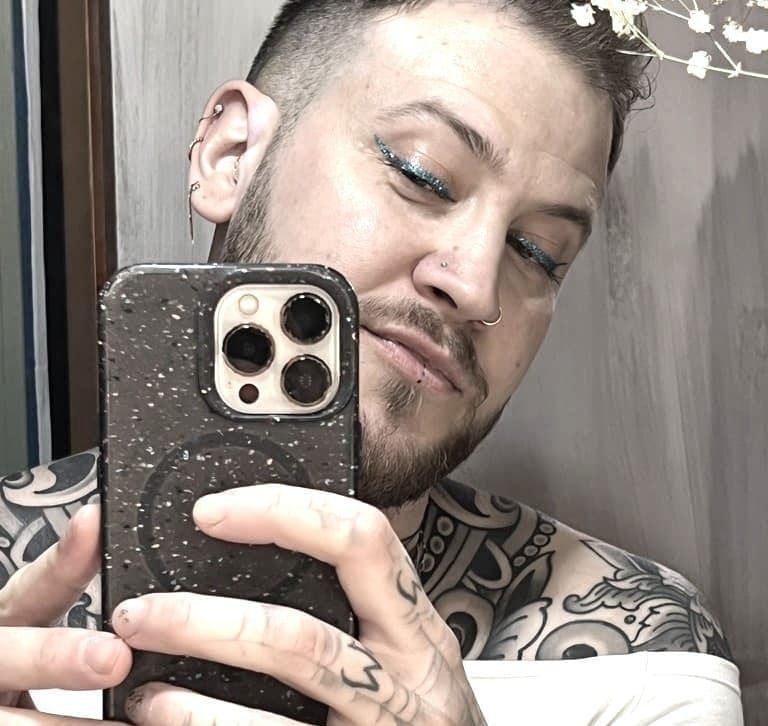 A man with a beard and blue-glitter cat-eye eyeliner takes a selfie in a mirror. He has black, flowery tattoos covering his bare shoulders.