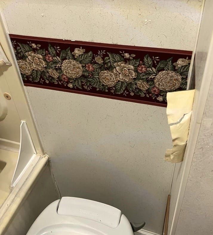 `A fraying strip of retro, burgundy-backed, horizontal floral wallpaper above a plastic toilet.