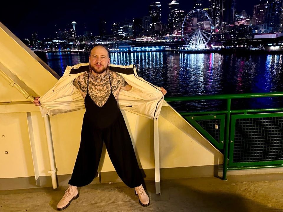A man stands on a ferry in front of the Seattle skyline at night. He is opening his white coat to show a strappy black bodysuit and his chest covered with black tattoos.