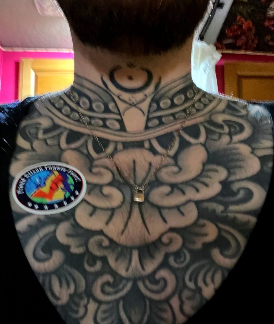 An image of a man’s chest, heavily tattooed with a black, deconstructed lotus, and a check-mark voting sticker on his right pec.