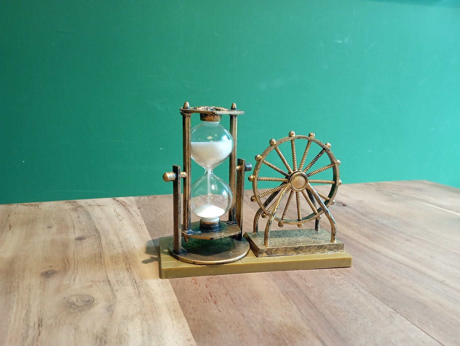 A small brass hourglass with white sand, mounted next to a decorative Ferris wheel on a wooden table in front of a green wall.