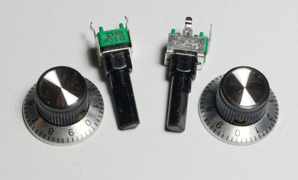 Two black and silver control knobs with numbered dial skirts from 0 to 9, lying beside two loose potentiometers on a white surface.