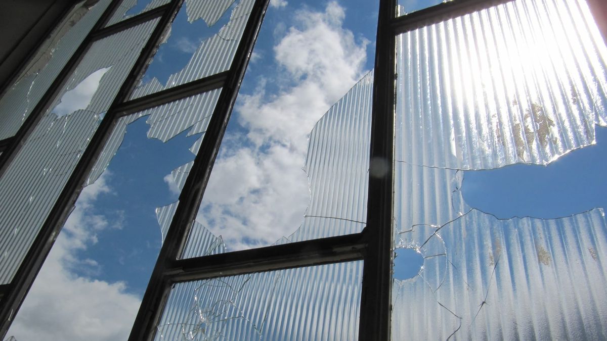A shattered industrial window with large jagged holes and cracked, ribbed glass panels, revealing a bright blue sky with scattered clouds behind the broken panes.