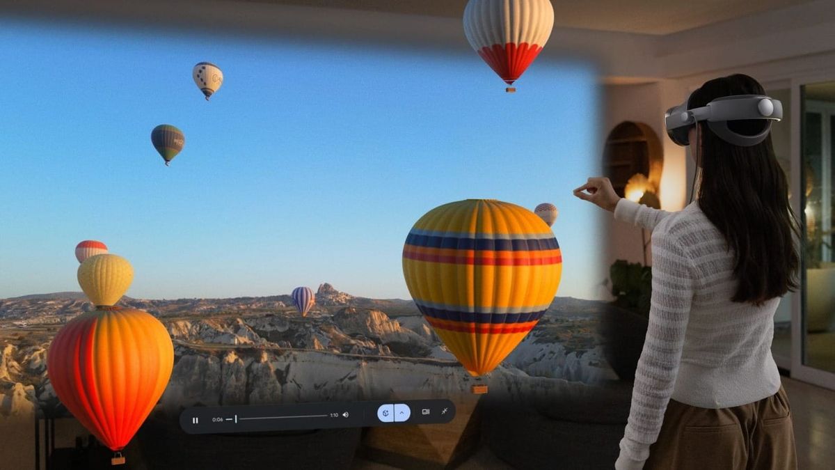 A woman wearing a mixed-reality headset stands in a living room, viewing a large immersive projection of colorful hot air balloons floating over a rocky landscape. 