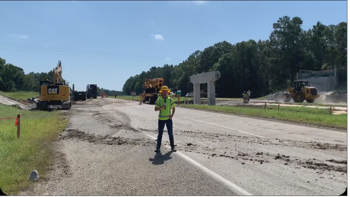 A man in a yellow reflective vest speaks into a microphone in front of a busy highway construction scene