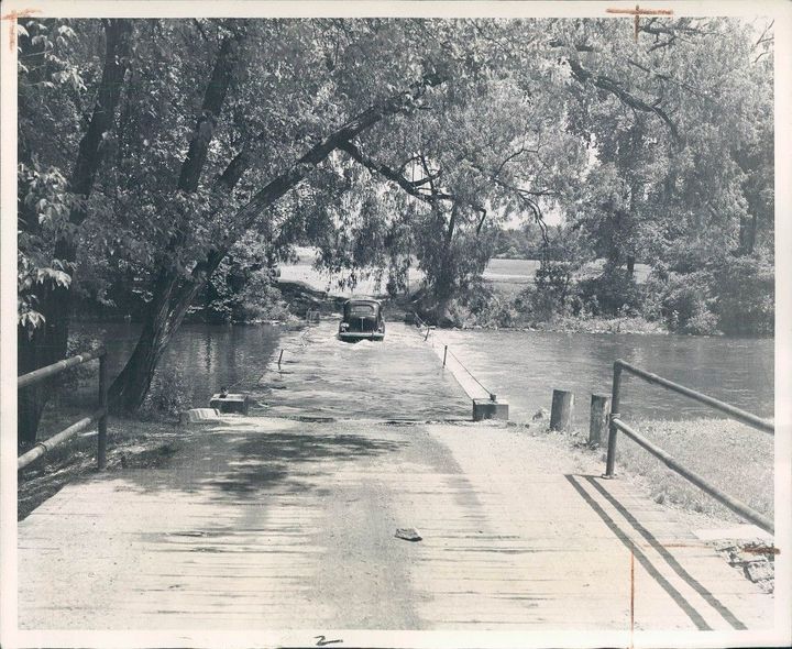 A black-and-white image of an old car driving across a sunken wooden bridge spanning a river