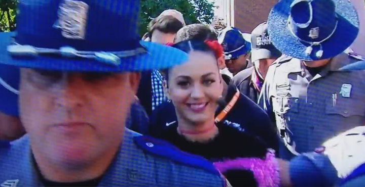 A broadcast image of pop star Katy Perry walking through a crowd escorted by many large policemen
