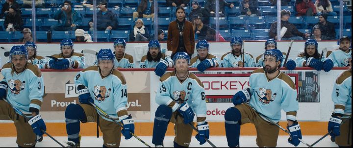 four hockey players in blue jerseys and brown pants kneel on the ice in front of the team bench