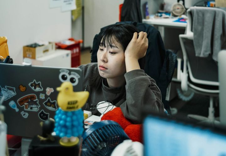 A female employee in a cluttered office looks boredly at her computer while resting her head on her hand